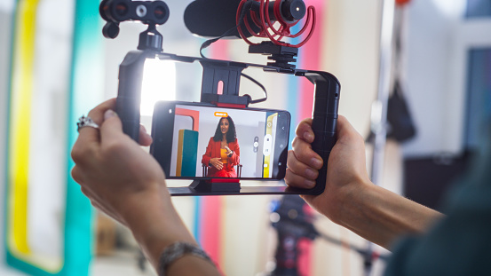 Young Female South Asian Host In Red Suite Engaging In Lively Discussion On Colorful Set, Captured On Smartphone And Professional Camera By Cinematographer For Live Broadcast Or Video Production.