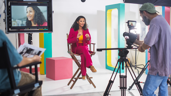 Young Hispanic Woman In A Vibrant Red Suit Speaks Animatedly On A Director's Chair, Surrounded By Colorful Set Design, With A Male Cinematographer Recording For A Lively Television Or Web Broadcast.
