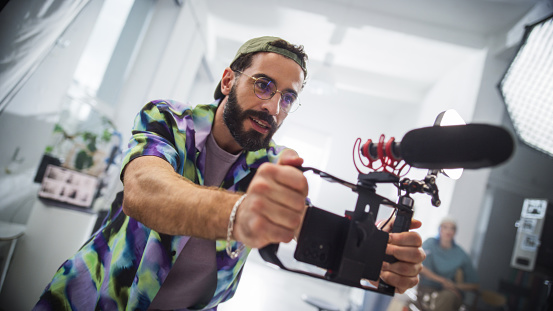 Young Latin Male Videographer With Beard, Wearing Colorful Shirt And Headband, Operates Camera With Microphone In Studio Setting, Capturing Creative Content For Advertising Or Movie Production.