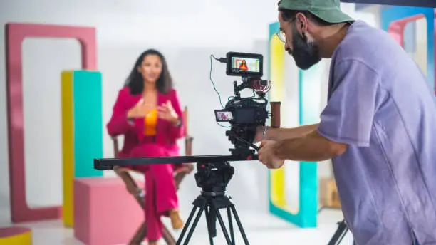 Young Male Cinematographer With Beard, Wearing Cap, Records Vibrant Female Host In Pink Suit During Lively Talk Show Set With Colorful Geometric Shapes, Enhancing Modern Video Production, Storytelling