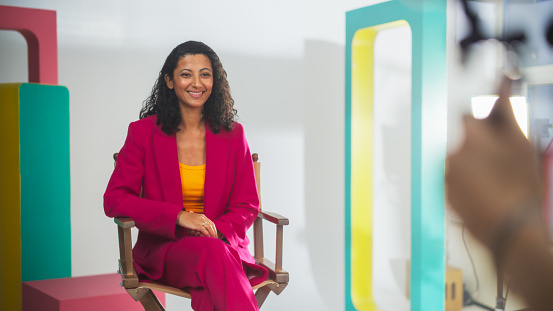 Young Middle Eastern Female Host In Vibrant Pink Blazer And Yellow Top, Seated On Director's Chair On Colorful Film Set, Smiling At Camera, With Blurred Videographer In Foreground.