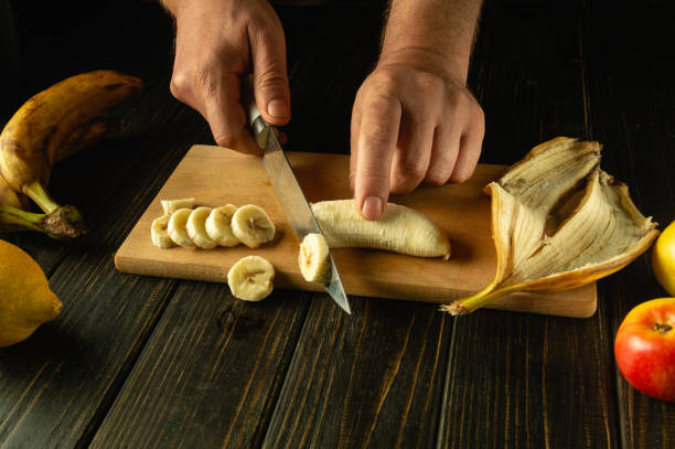 istock Preparing a fruit dish on the kitchen table. Chef hands using a knife to slice a banana on a wooden cutting board 2149807283