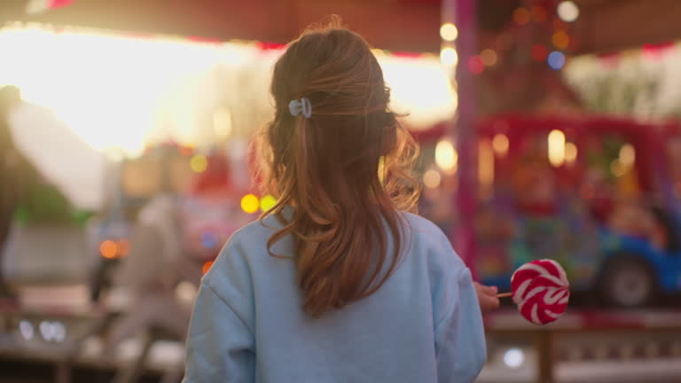 A little lovely girl holds a large striped lollipop in her hand and looks at a glowing attraction in an amusement park at sunset