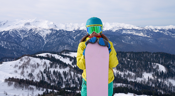 A snowboarder with a beaming smile hugs her board, standing before a sweeping view of alpine peaks, embodying the joy of winter sports.