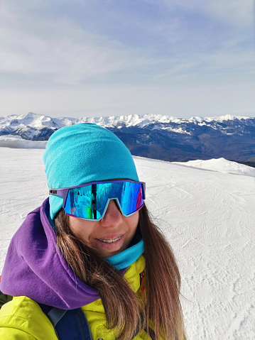 Close-up selfie of a smiling skier with reflective goggles, showcasing a breathtaking view of distant snow-capped mountains under a serene sky.