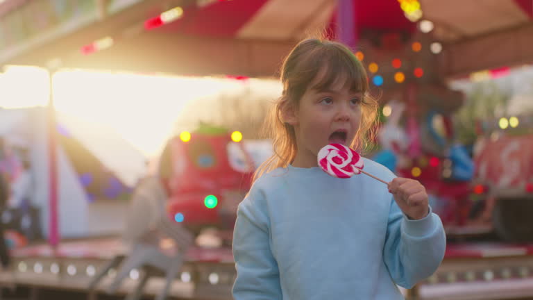 A small beautiful girl holds a large striped lollipop in her hand and eats it against the backdrop of glowing rides in an amusement park