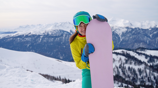A young snowboarder hugs her board, smiling against the waning light of the dusk sky over snowy mountains, ready for the next day's thrills.