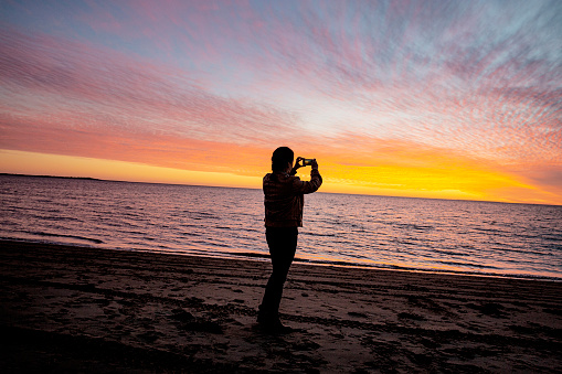 A woman taking a photograph of a beautiful vibrant sunset with he cell phone, whilst on a beach at the edge of the ocean in the state of Sinoloa in Mexico