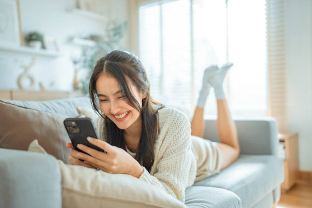 istock Happy young asian woman lady browsing surfing wireless internet on mobile phone while sitting a sofa couch in living room at home apartment, Shopping online via website 2149818112