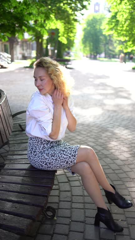 beautiful middle age woman in 70s, 80s style clothes on a bench in a sunlit avenue