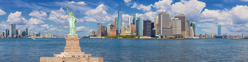Statue of Liberty and New York City Skyline with Jersey City, NJ, Manhattan Financial District, Battery Park, World Trade Center, Staten Island Ferry, Brooklyn Bridge, Manhattan Bridge, Blue Sky with Puffy Clouds and Water of New York Harbor. High Resolution Stitched Panoramic image with 4:1 image aspect ratio. This image was downsized to 50MP. Original image resolution is 116MP or 21,572 x 5,393 px. Canon EOS 6D Full Frame Sensor Camera and Canon EF 85mm f/1.8 USM Prime Lens.