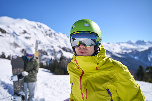 In the foreground, a skier with reflective goggles and vibrant attire stands poised against a breathtaking alpine backdrop, snowboarder in tow.