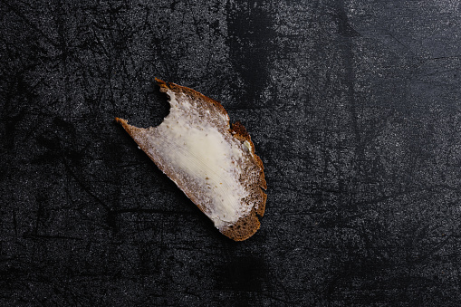 A bitten bread is unevenly spread with butter. It is placed on a dark, stone-patterned background. The Close-up reveals a slice of a bitten farmhouse bread smudged with German butter. The shot is captured in an overhead view. Still life of bitten Slice Farmhouse Bread Directly Above Shot.