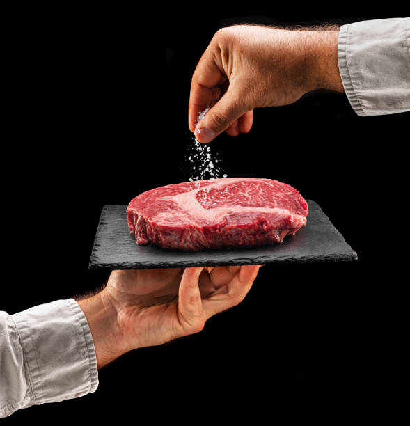Chef is salting or seasoning raw ribeye steak laying on graphite serving board. Black background.