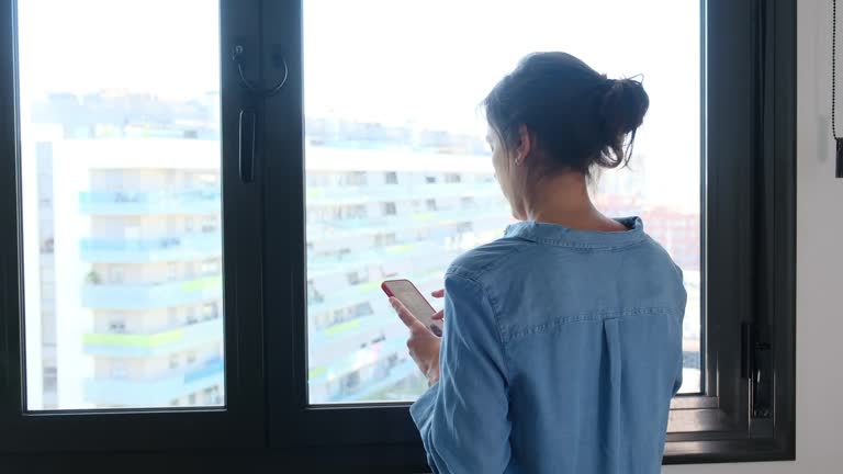 Woman with her back in front of a window in a big city typing with mobile chat reviewing web application, internet shopping and mobile applications