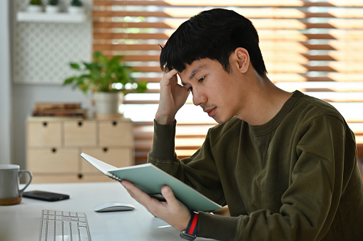 Young Asian man working at home office, feel tried while reading a book.
