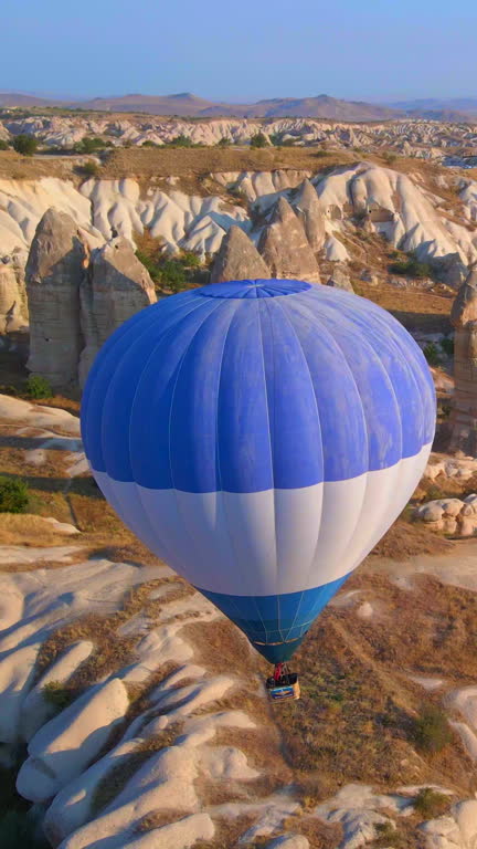 Vertical aerial video. Captured against the canvas of the Cappadocian sky, this video showcases the enchanting spectacle of a hot air balloon festival. One blue ballon gracefully glides among distinctly shaped rocks of the Love Valley in Gureme in