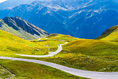 Panoramic View of Winding High Alpine Road Grossglockner Amidst Lush Green Hills, Rugged Mountains, and Blue Sky with Clouds