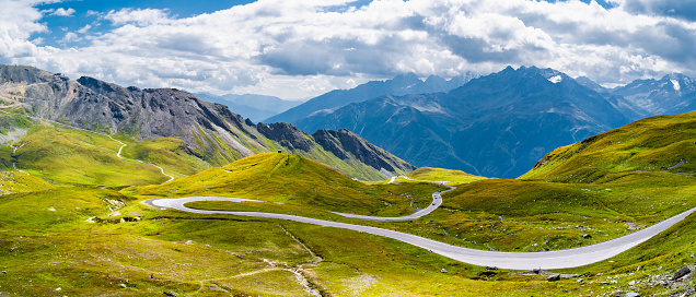 Winding road Grossglockner in Austria amidst lush green hills and rugged mountains in the background under a blue sky adorned with fluffy clouds.