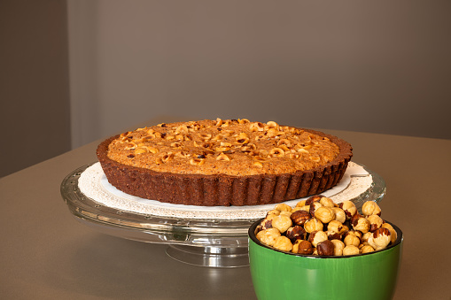 A homemade chocolate tart with a delectable hazelnut cream is complemented by a bowl of roasted hazelnuts, presenting a rich and nutty culinary delight