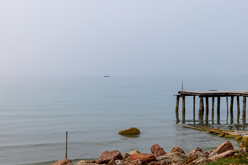 A weathered pier juts into the placid waters of Lake Garda, while a solitary boat on the horizon drifts through the fog on a quiet Italian morning