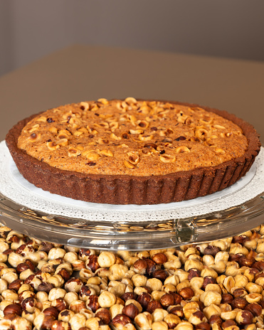 A freshly baked chocolate hazelnut tart sits elegantly on a glass stand, its surface generously adorned with golden toasted hazelnuts