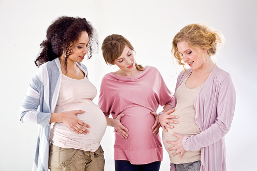 Three happy and proud, pregnant young adult women standing against white background and holding their pregnant bellies.
