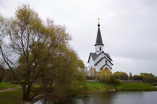 A church with white walls and a dark roof situated by the lakeside, surrounded by green trees and grass under a cloudy sky.