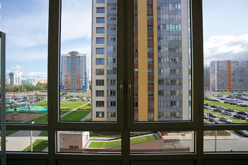 View from a window onto an urban landscape with numerous high-rise buildings of various heights and colors. In the foreground, there are window frames, below is a parking lot full of cars and a green area with a playground. The sky is clear with a few clouds.