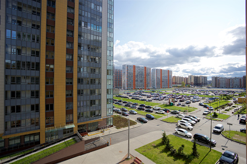 View of an urban landscape with tall residential buildings of various colors, a vast parking lot full of cars, and green plantings on a bright sunny day with a cloudy sky.
