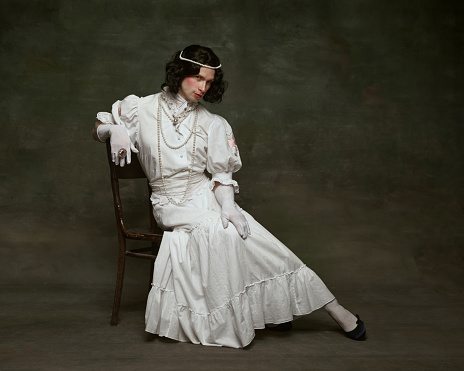 Pensive man wearing period gown and wig, adorned with pearls and seated on wooden chair against vintage studio background. Concept of self-expression, comparisons of eras, freedom, human rights.