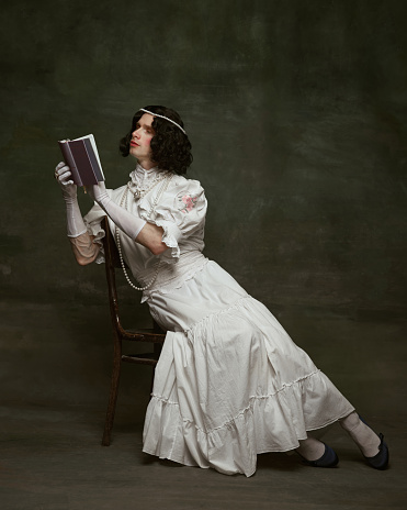 Young attractive ma dressed as medieval woman sitting on wooden chair and reading book against vintage studio background. Concept of self-expression, comparisons of eras, freedom, human rights.