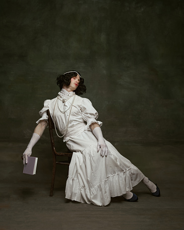 Man in white dress and black wig reclining on chair with eyes closed, holding book against vintage studio background. Concept of self-expression, comparisons of eras, freedom, human rights.