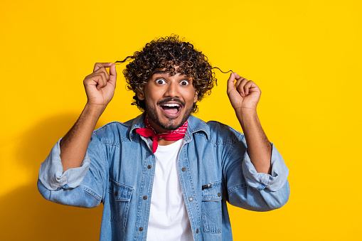 Photo portrait of handsome young guy hold excited curly strands dressed stylish jeans garment red scarf isolated on yellow color background.