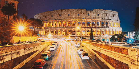 Rome, Italy. Colosseum Also Known As Flavian Amphitheatre In Evening Or Night Time. Night Traffic Light Trails Near Famous World Landmark. Amazing Night Starry Sky Background. Bright Blue Night Sky.