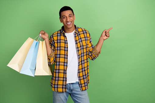 Photo of handsome guy hold store bags indicate finger empty space offer isolated on green color background.