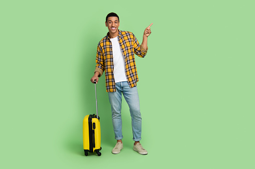 Full length portrait of nice person hold suitcase indicate finger empty space ad isolated on green color background.