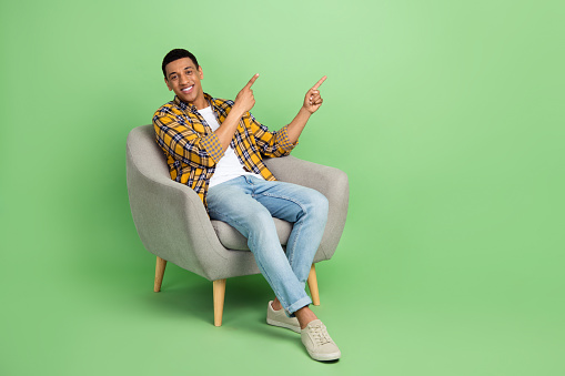 Full length portrait of positive guy sit cozy chair indicate fingers empty space offer isolated on green color background.