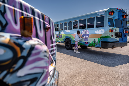 Mid adult Iranian woman artist painting  exterior mural on party bus, being converted from traditional school bus.  She is dressed in casual work clothes. Exterior of old school bus.