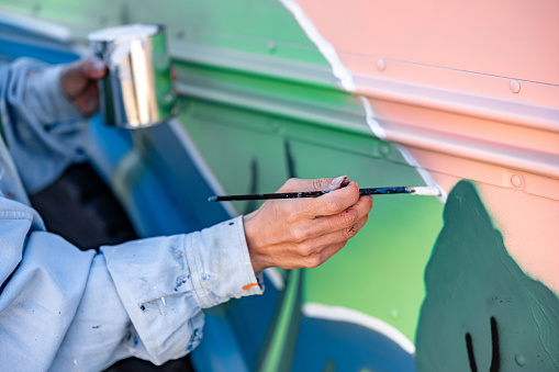 Closeup of hands of Mid adult Iranian woman artist painting  mural on  party bus.  She is dressed in casual work clothes. Exterior of old school bus.