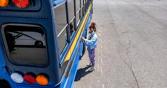 Mid adult Iranian woman artist painting  exterior mural on party bus, being converted from traditional school bus.  She is dressed in casual work clothes. Exterior of old school bus, drone point of view.