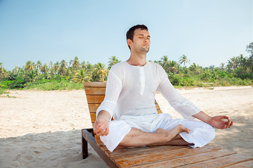A young adult man peacefully relaxing and meditating on a sunny tropical beach, surrounded by serene beauty. Ideal for wellness, travel, and relaxation concepts.