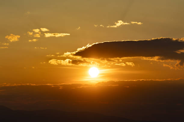 The golden sun dips below the clouds, casting a warm light over the silhouette of a mountain range.