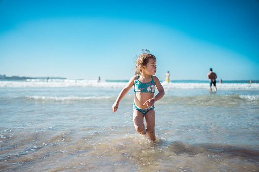 An adorable three year old multiracial girl stands knee deep in the ocean, playing and splashing during a family vacation in Australia.