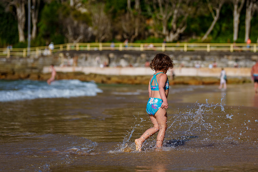 An adorable three year old multiracial girl stands ankle deep in the ocean, playing and splashing during a family vacation in Australia.