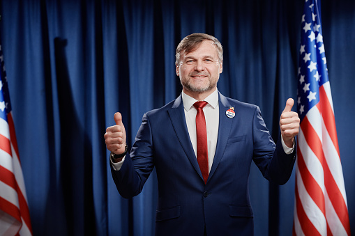 Successful mature male politician in formalwear showing thumbs up and looking at camera with smile while standing against American flag