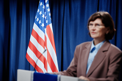 American flag on platform with microphone and defocused mature female politician in formalwear standing in front of public during speech