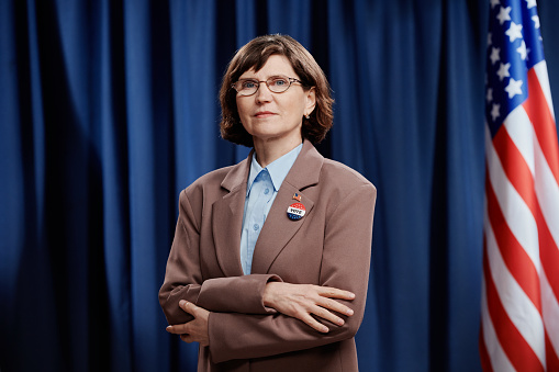 Mature female politician in eyeglasses and formal suit keeping her arms crossed by chest and looking at you after press conference