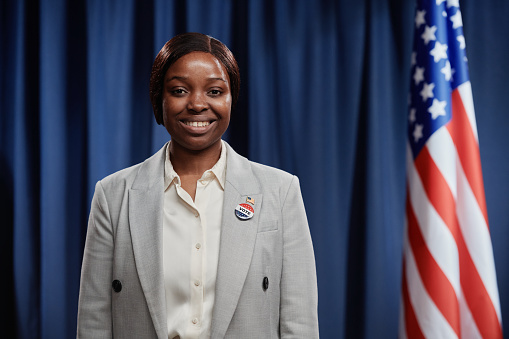 Beautiful African politician in a red suit posing on camera with a smile before  the speech, standing against the blue background with American flags