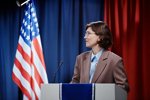 Mature brunette female politician looking aside while standing by platform, speaking in microphone and making presentation